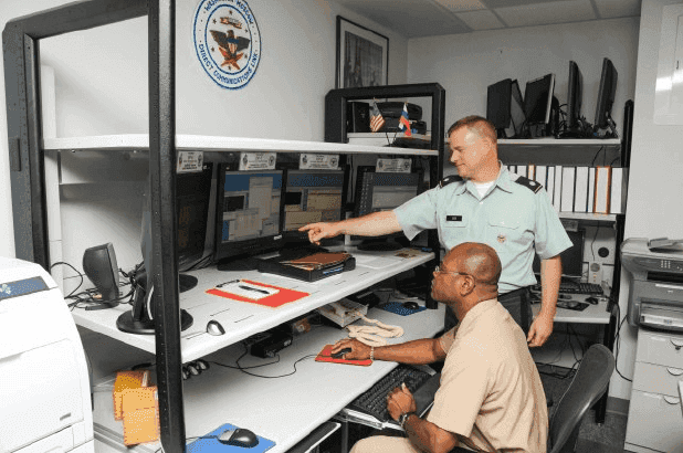 Two military officers working on a computer sitting on a desk in front of them. One officer is point at the screen and the other officer is using the mouse to select something on screen.